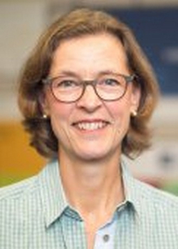 Eine lächelnde Frau mittleren Alters mit kurzen braunen Haaren und Brille, die direkt in die Kamera blickt.