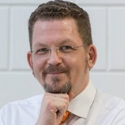 Mann mit Brille und Bart, der nachdenklich lächelt und die Hand am Kinn hält.
