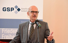 Ein glatzköpfiger Mann mit Brille und Schnurrbart spricht ins Mikrofon vor einem Banner mit "GSP", "Frieden" und "Freiheit".