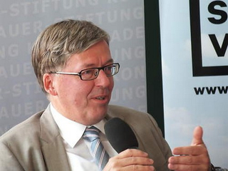 Ein Mann in Anzug und Brille spricht in ein Mikrofon und gestikuliert, mit Event-Bannern im Hintergrund.